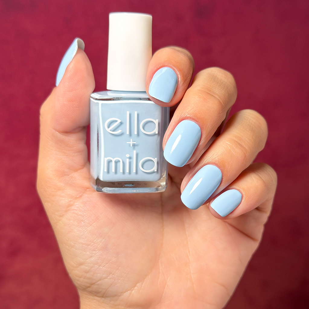 A hand with light blue painted nails holds a matching bottle of Ella + Mila nail polish.