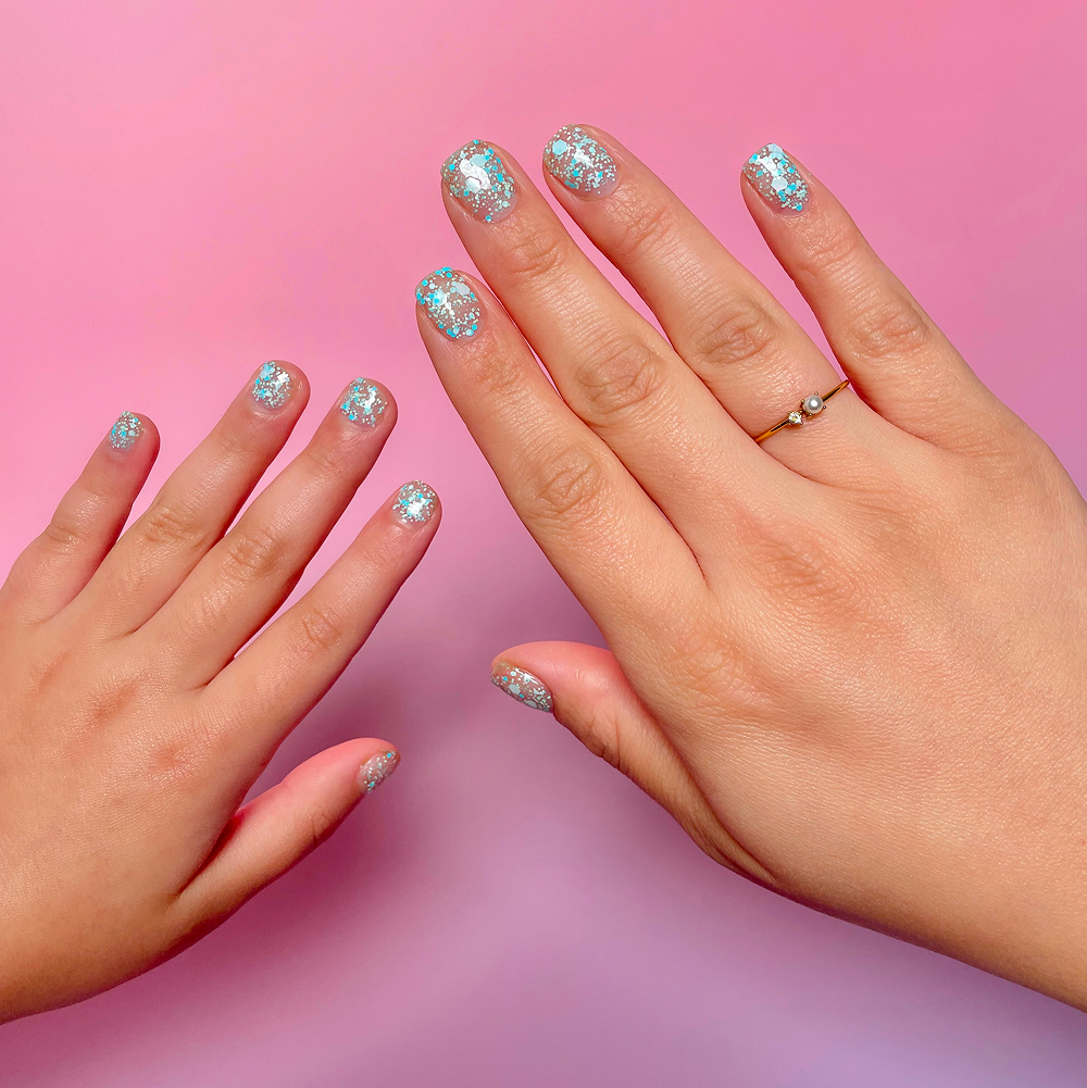 An adult's and a child's hands with matching blue and silver glitter nail polish on a pink background.