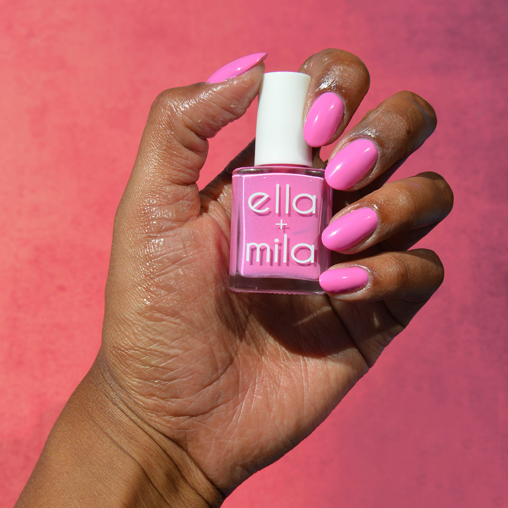 A hand with a bright pink manicure holds a matching bottle of Ella + Mila nail polish.