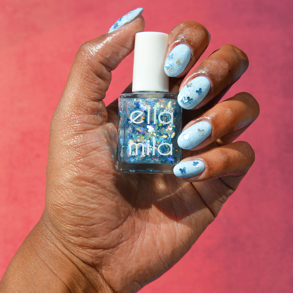 A hand with a light blue butterfly manicure holding a bottle of Ella + Mila glitter nail polish.