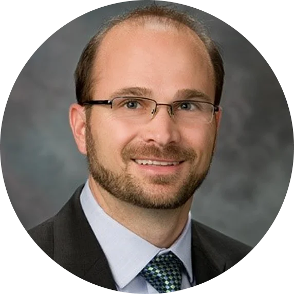 A circular headshot of a smiling man with glasses and a beard, wearing a suit and tie.