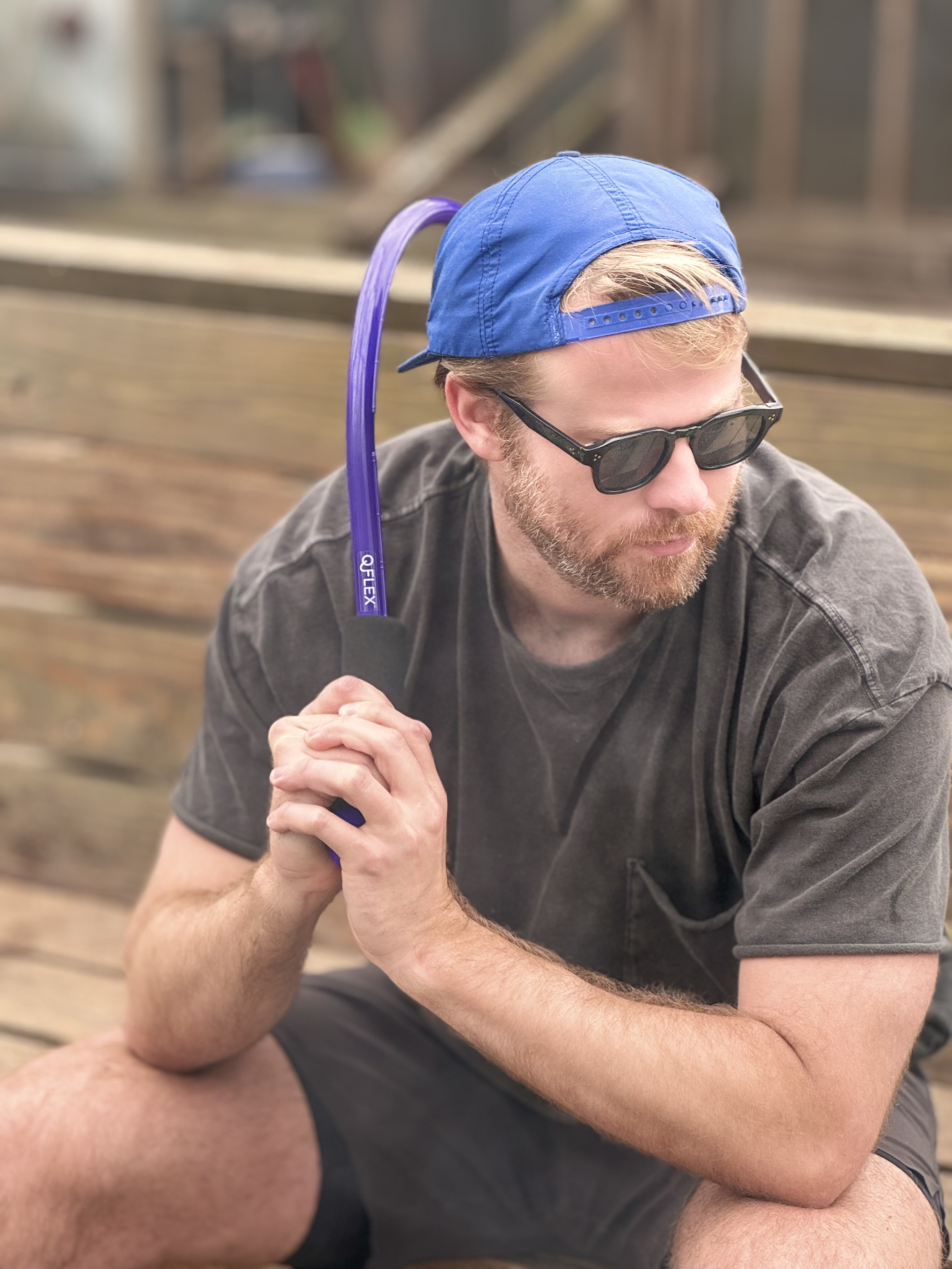 Man in sunglasses and blue cap holding a purple object, sitting on a wooden bench.