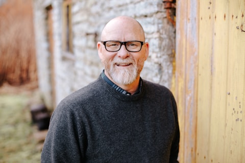 Man with glasses and beard smiling, standing outside near a stone building.