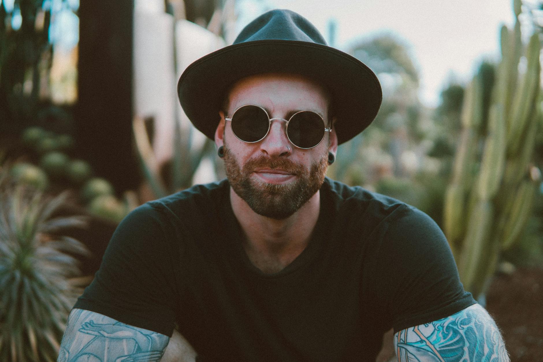 Man with sunglasses and hat, sitting outdoors with plants in background.