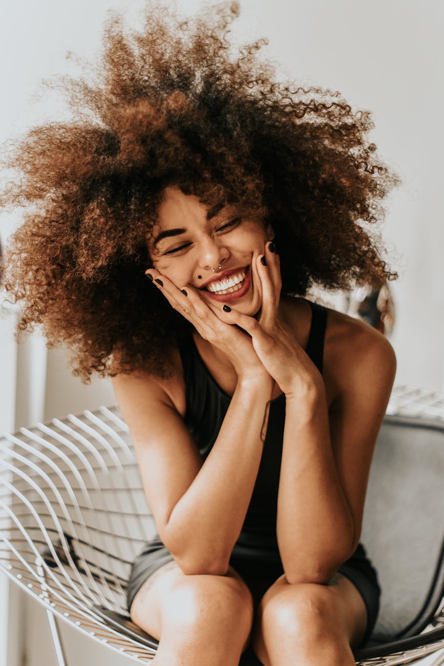 Smiling person with curly hair sitting on a chair, hands on cheeks.