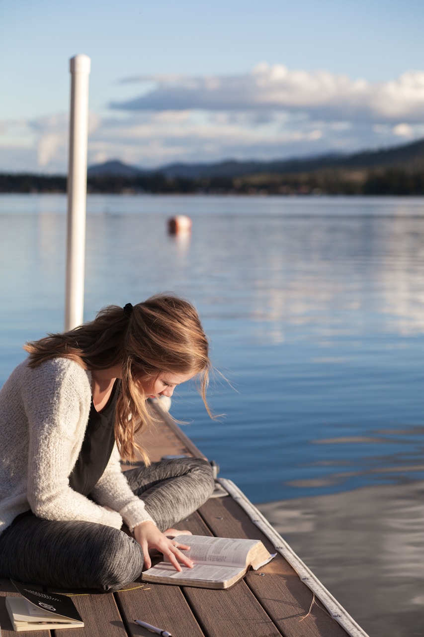 Person reading a book on a dock by a lake.
