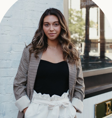 A woman with long brown hair in a blazer and white pants stands against a brick wall.
