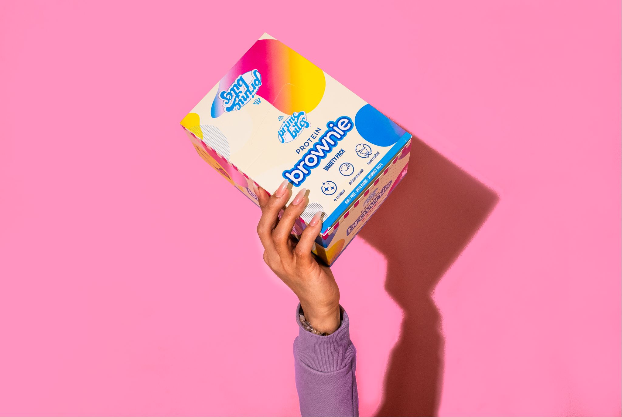 Hand holding a colorful box labeled 'protein brownie' against a pink background.