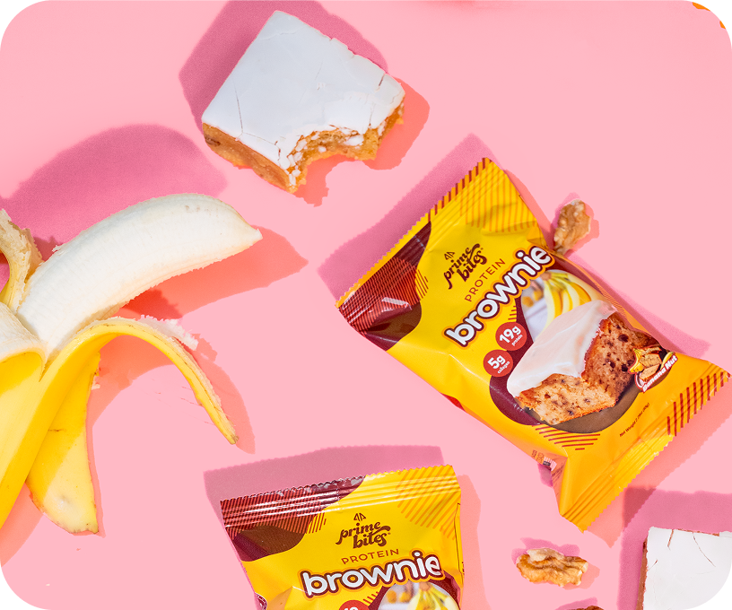 Banana, protein brownie packages, and brownie square on pink background.