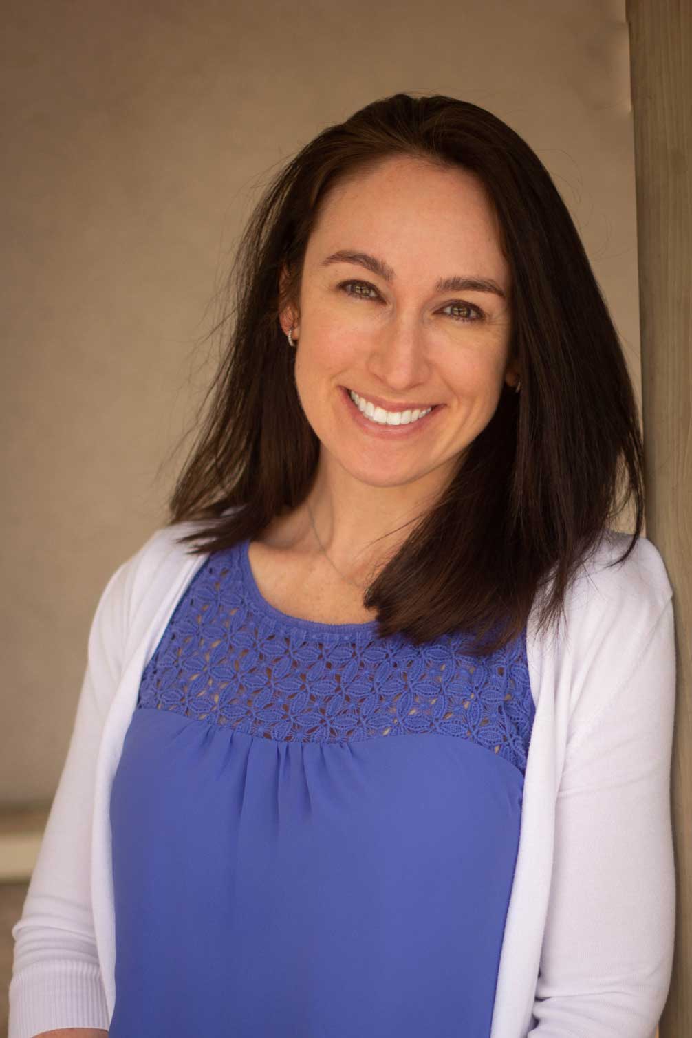 A smiling woman with long brown hair, wearing a blue top and white cardigan, leaning against a wall.
