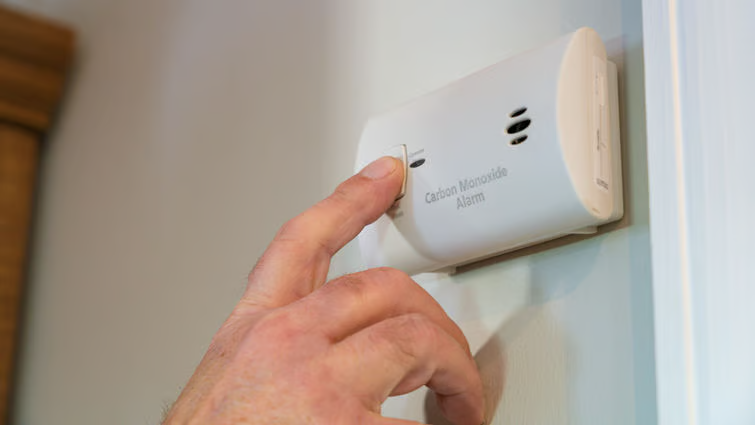 A person's hand pressing the test button on a white carbon monoxide alarm mounted on a wall.