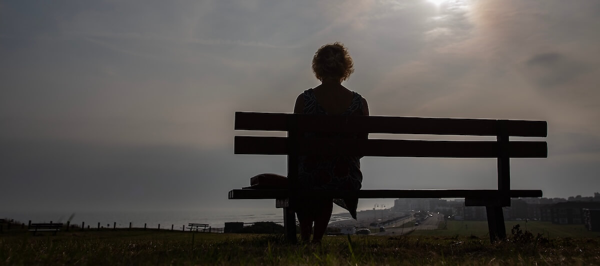 A silhouetted person sits on a bench overlooking a hazy coastal town and the sea.