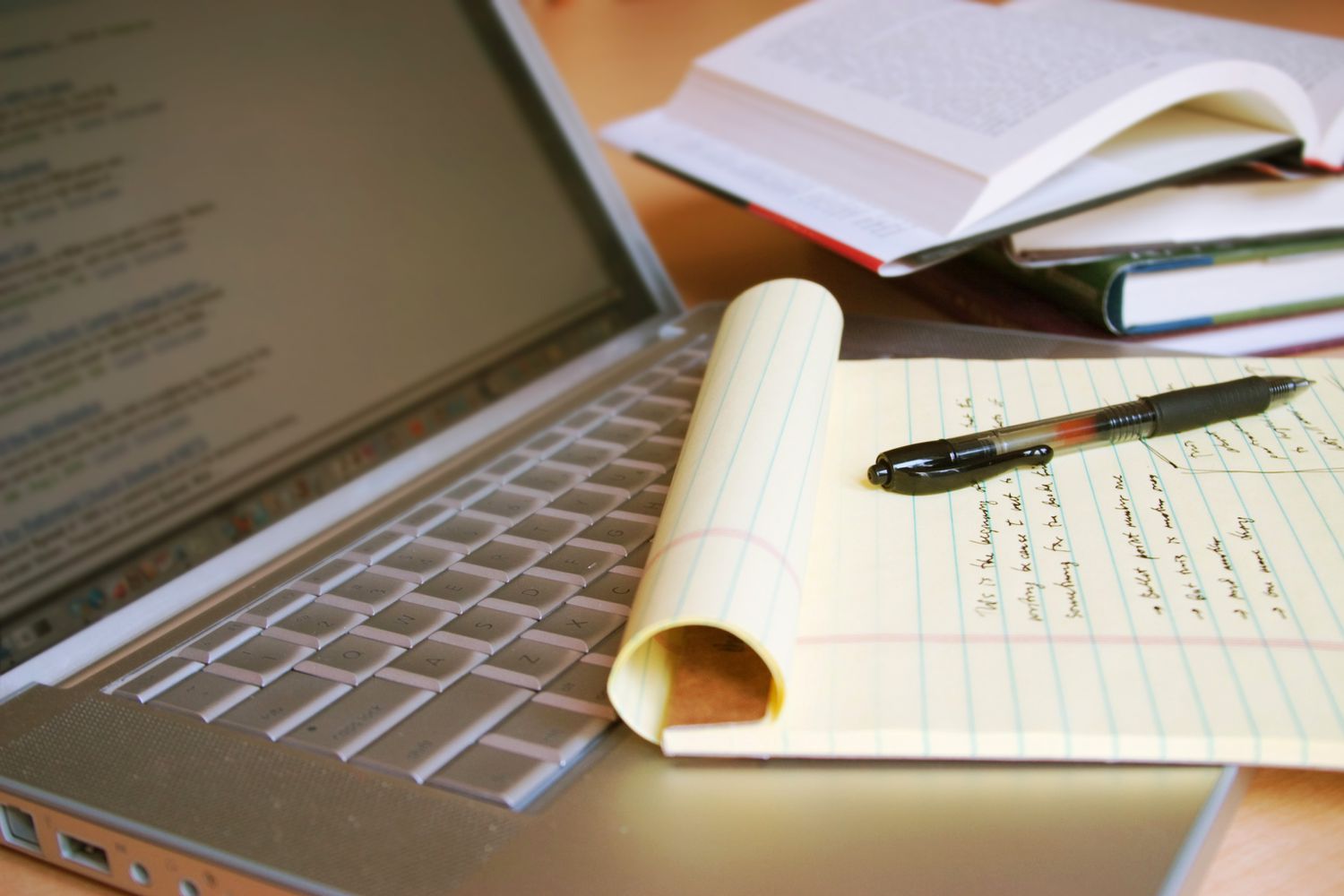 A laptop on a desk with a yellow notepad, a pen, and books in the background.
