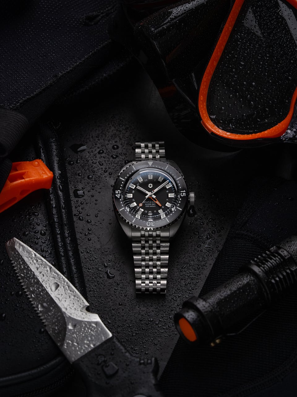 Silver watch surrounded by diving gear and knife on a wet surface.