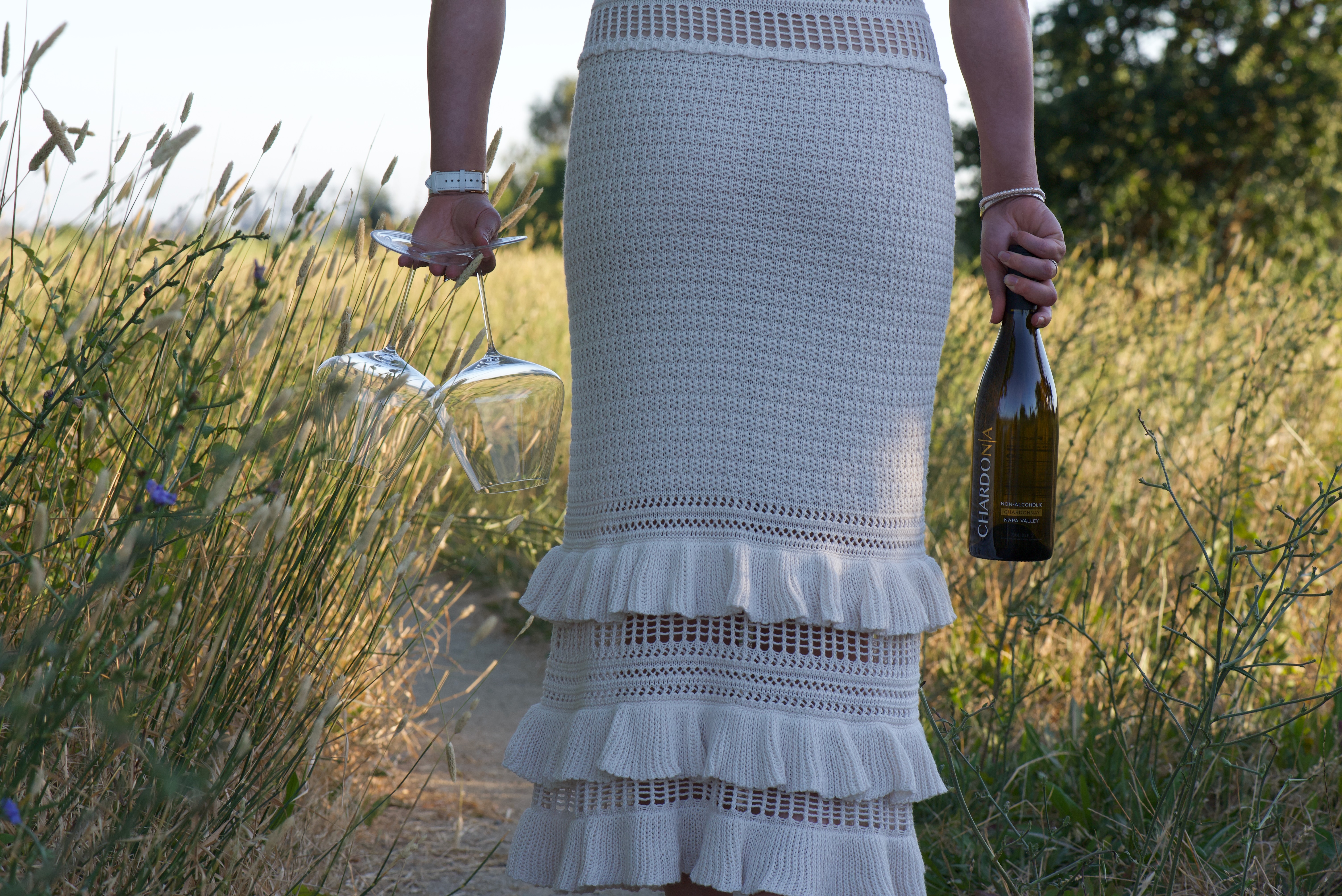 A person in a white skirt holds a bottle of Chardonnay and two wine glasses in a grassy field.