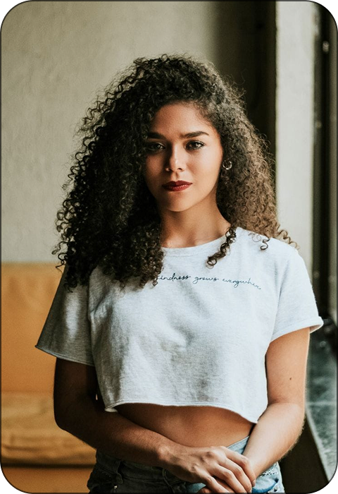Person with curly hair wearing a cropped white shirt, standing indoors.