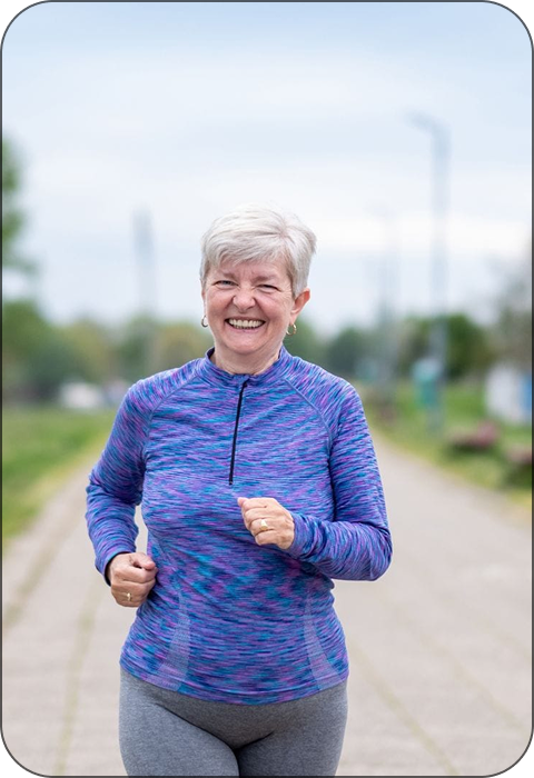 Elderly woman jogging on a path, wearing a purple and blue top.