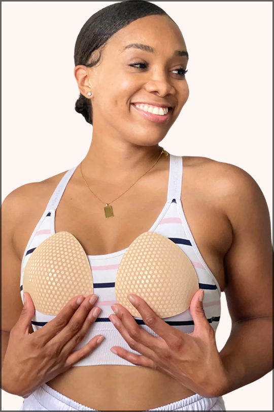 Smiling woman in striped sports bra holding beige Honey Cloudz triangle bra inserts, lightweight and swim-ready, perfect for halter tops, sports bras, and post-lumpectomy wear.