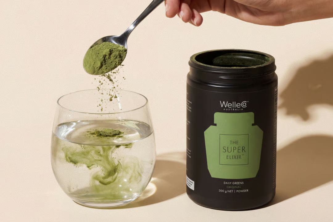 A hand pours a green smoothie from a blender into a glass next to a Super Elixir container.