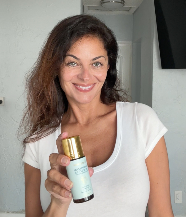 A smiling woman in a white t-shirt holds up a small bottle of frankincense and castor oil.