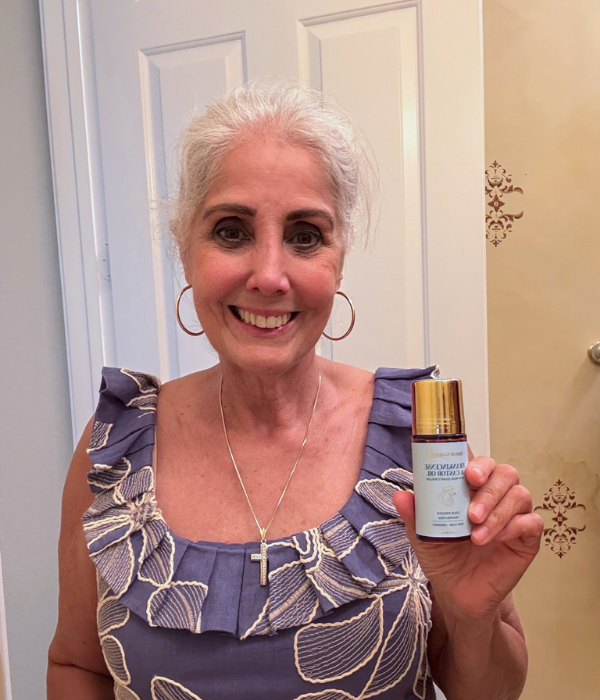 A smiling woman with white hair and a blue floral top holds up a small bottle.