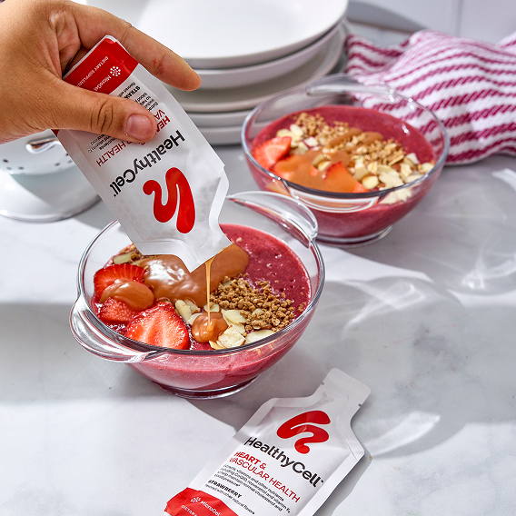 Hand pouring supplement into a smoothie bowl with strawberries and granola.