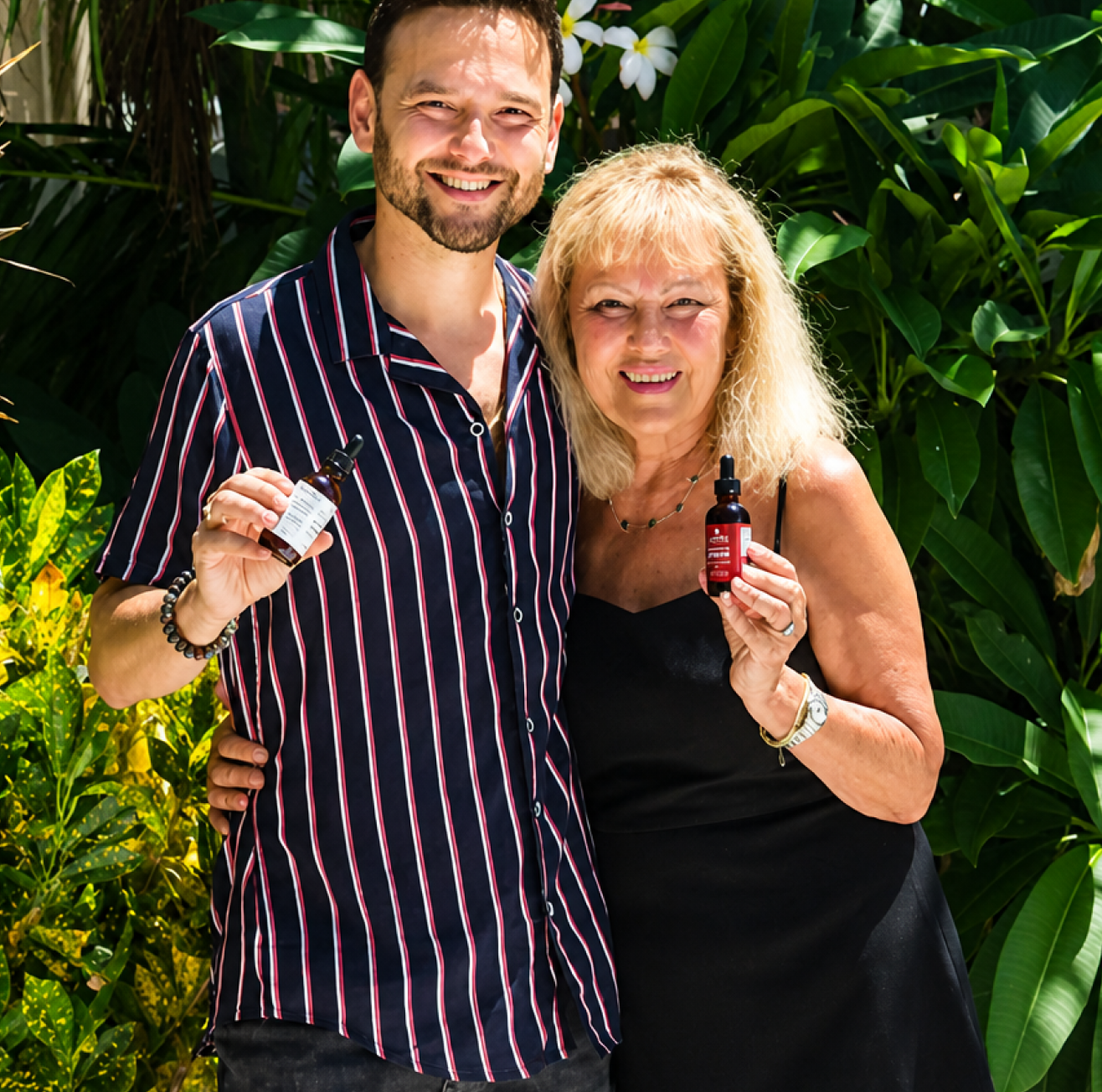 A smiling man and woman pose outdoors in front of plants, each holding a small dropper bottle.