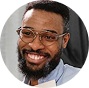 A close-up, circular-cropped photo of a smiling Black man with glasses and a beard.