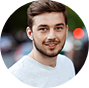 A circular headshot of a smiling young man with a beard, wearing a grey t-shirt, with a red dot.