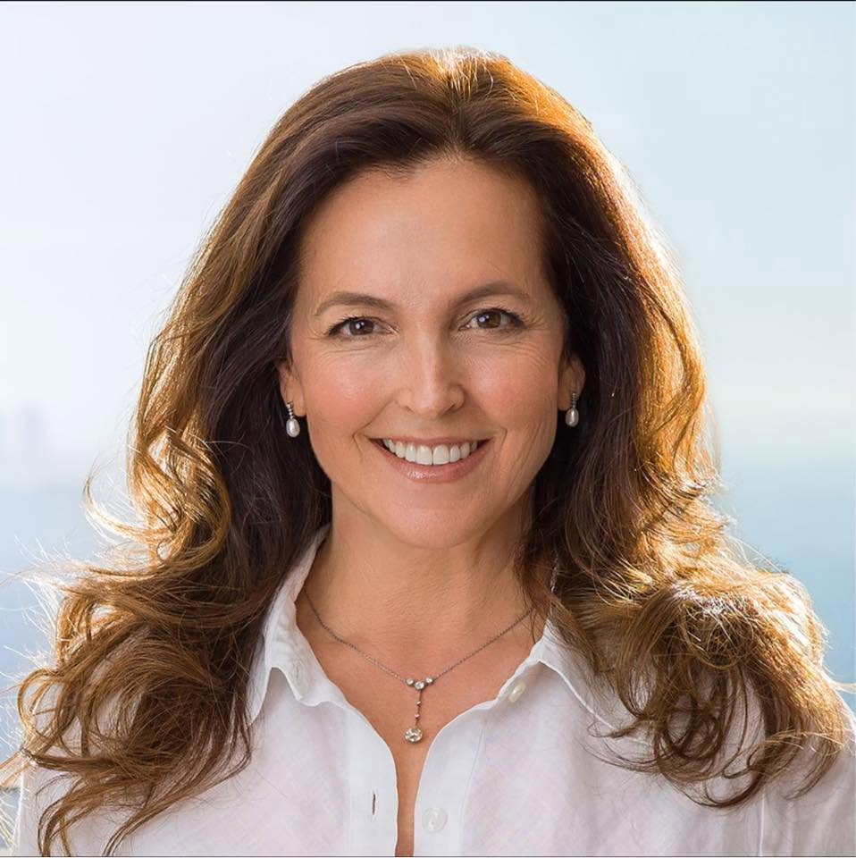 A smiling woman with long brown hair, wearing a white collared shirt, pearl earrings, and a necklace.
