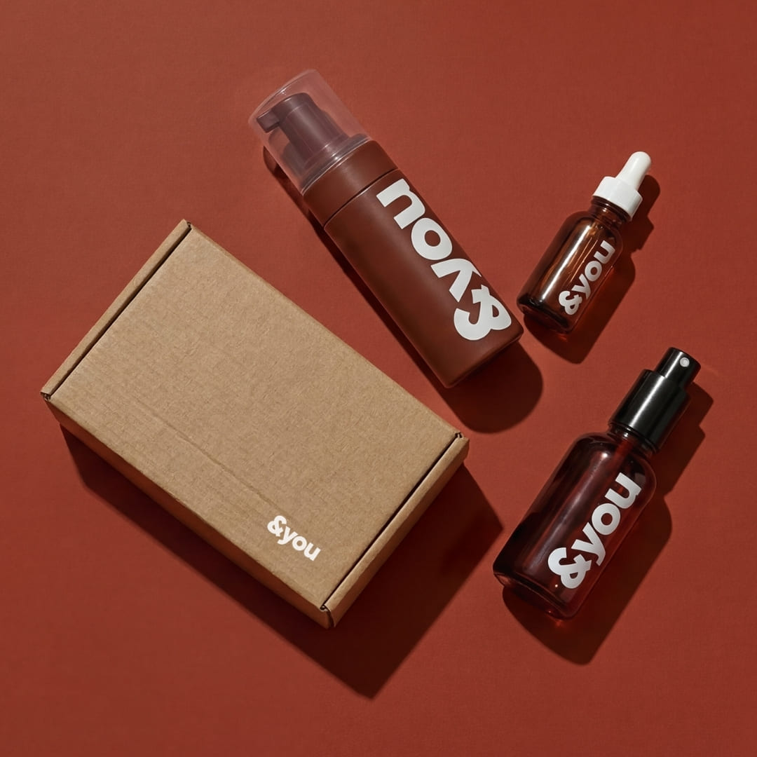Three '&you' branded cosmetic bottles and a small cardboard box on a reddish-brown background.