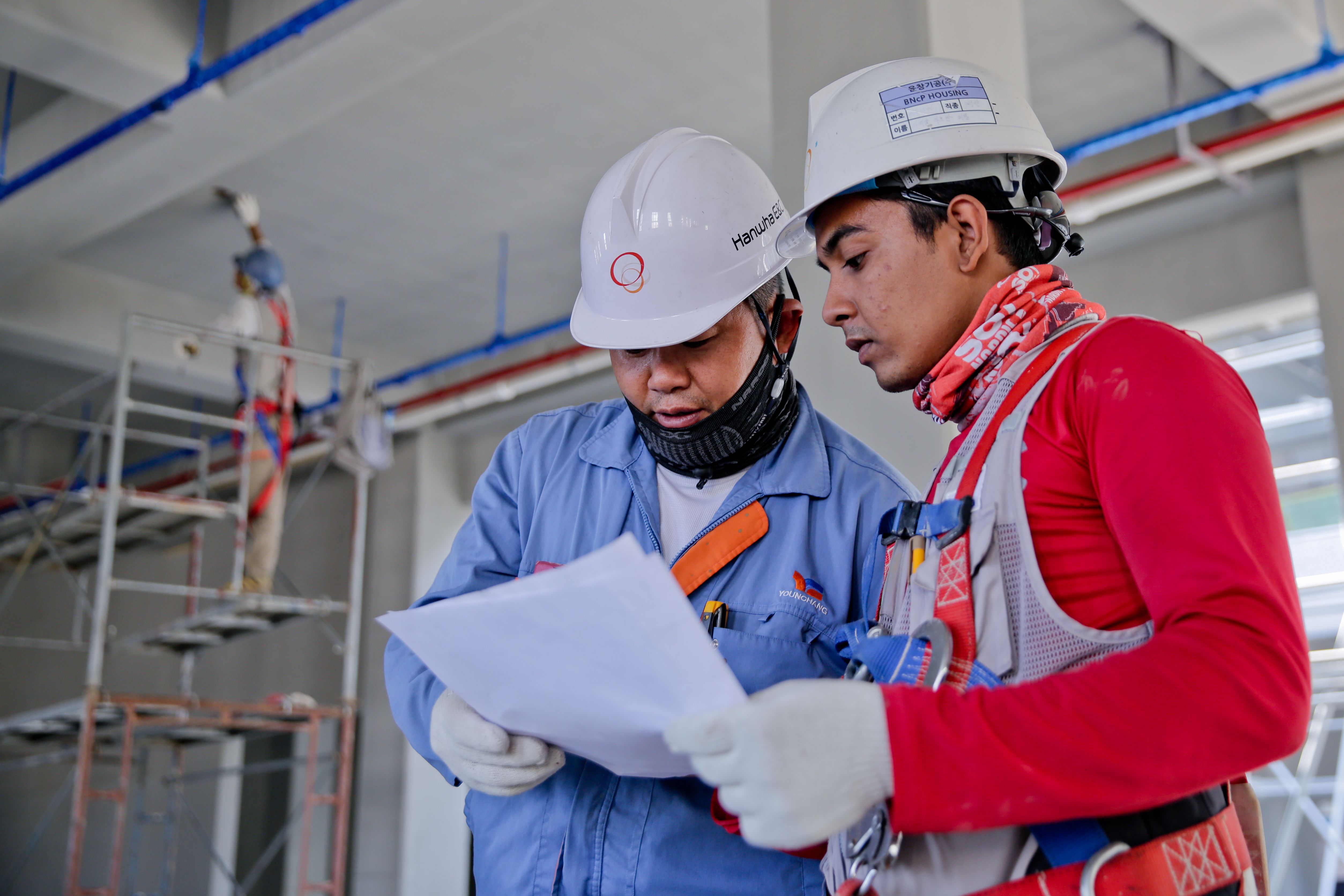 Two construction workers in hard hats review a document on a worksite with another worker on scaffolding behind them.