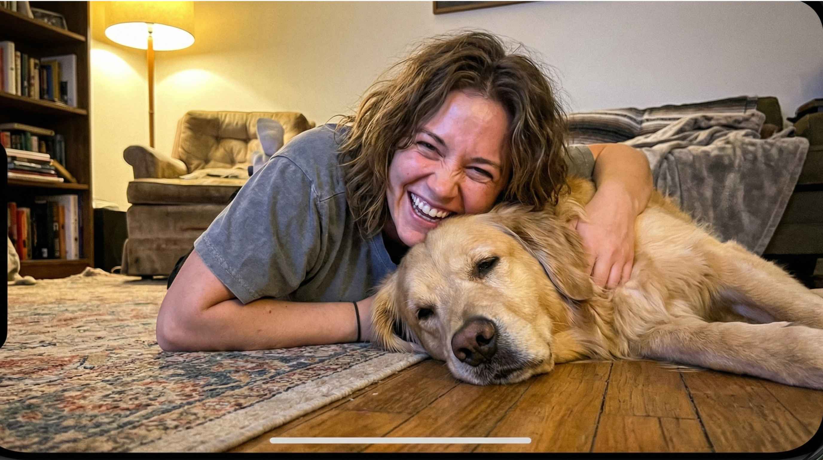 A laughing woman with curly hair hugs a golden retriever as they lie on the floor.