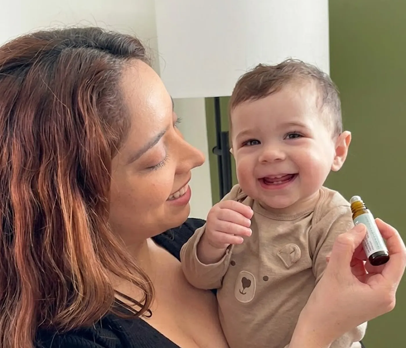 A smiling woman holds a laughing baby and a small rollerball bottle of essential oil.