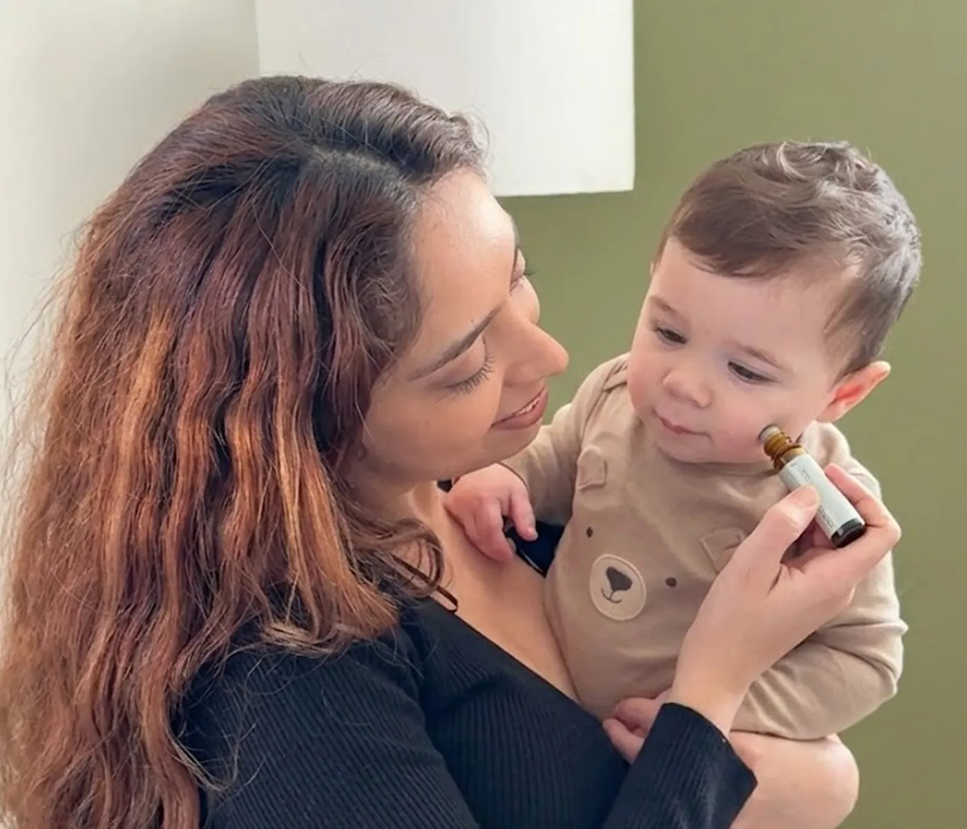 A woman holds a baby while applying a rollerball bottle to the baby's cheek.