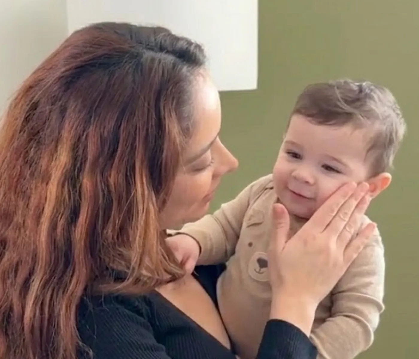 A woman with long hair holds a smiling baby and gently caresses its cheek.