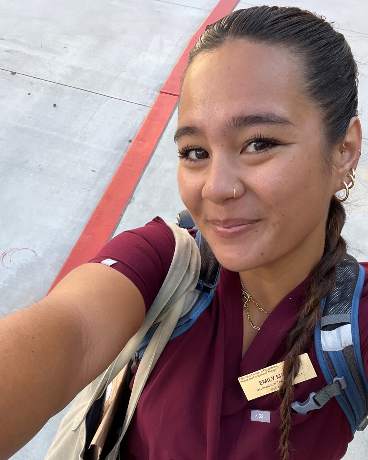 A woman in maroon scrubs and a backpack smiles while taking a selfie outdoors.