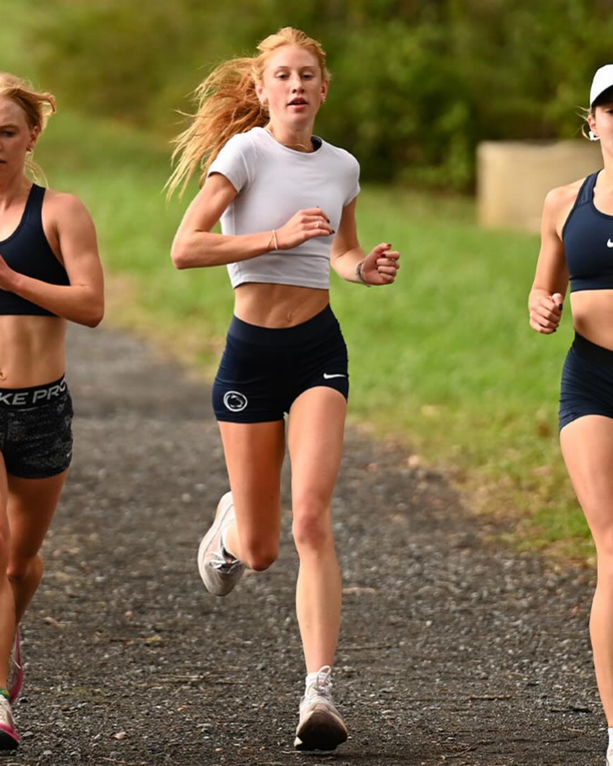 Three women in athletic gear run along a gravel path outdoors.