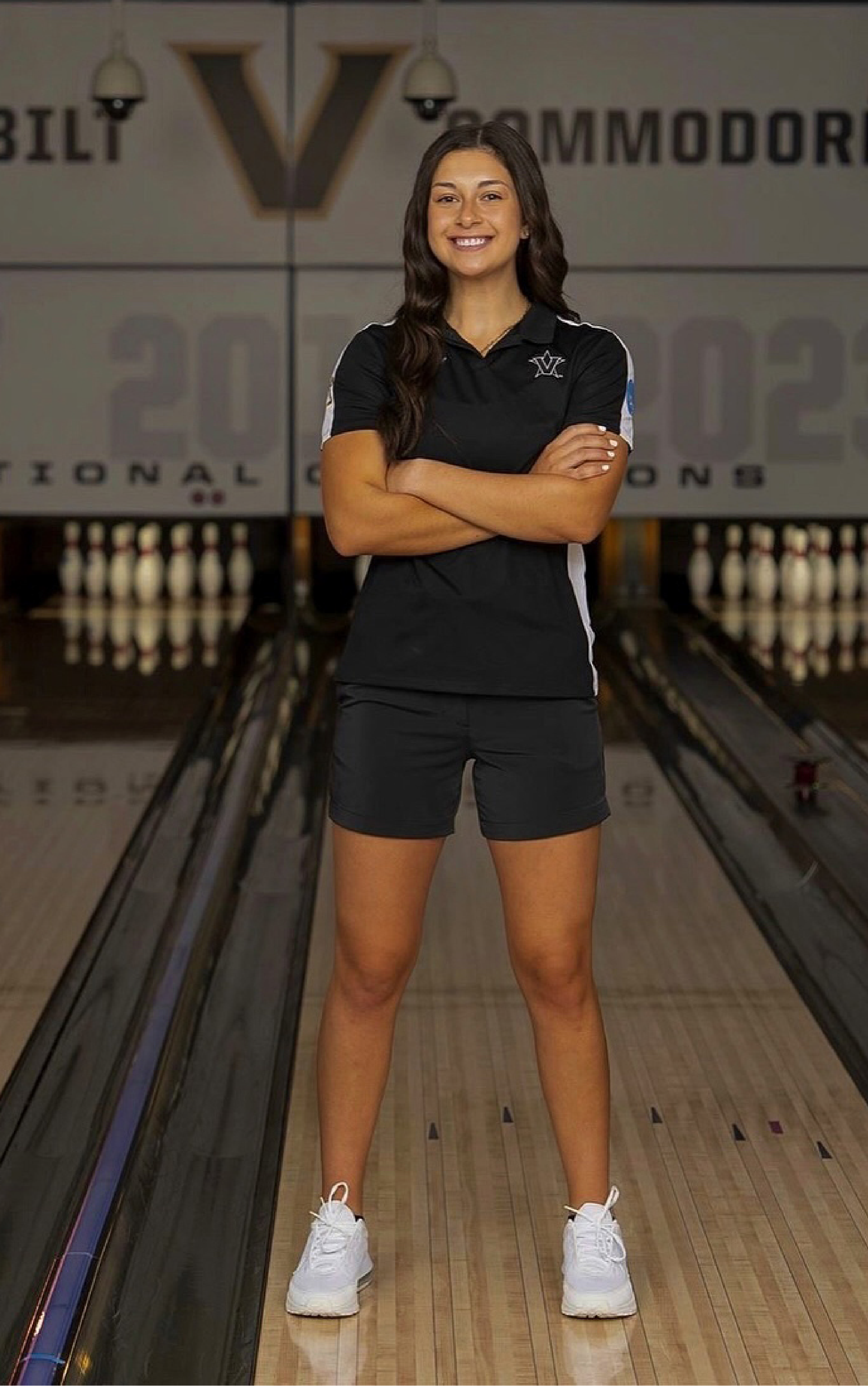A young woman in a Vanderbilt polo shirt and shorts stands smiling in a bowling alley with arms crossed.