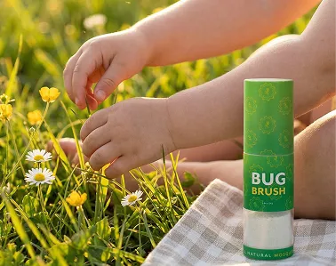 A child's hands reach into a grassy field next to a Bug Brush container on a blanket.
