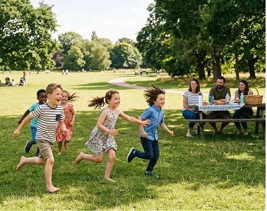 A group of children run laughing across a grassy park while adults watch from a picnic table.