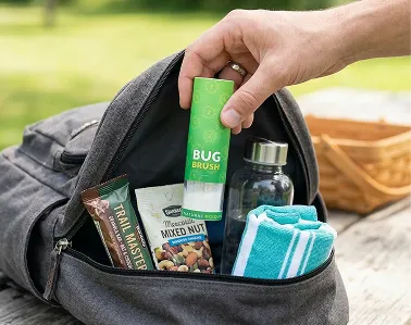 A hand places a green bug brush into a backpack with snacks and a water bottle.