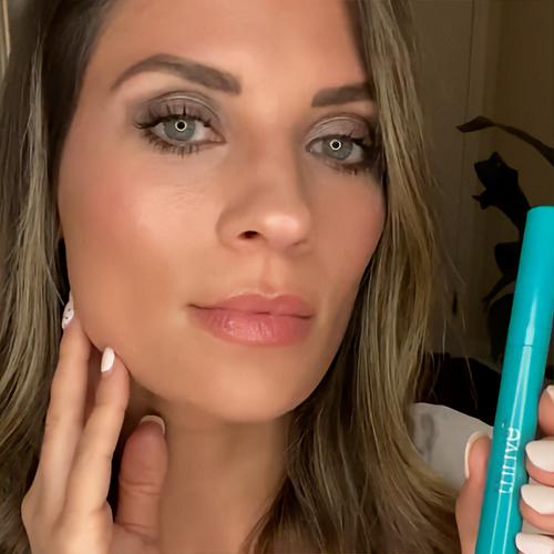 Woman holding a turquoise mascara tube, with a neutral expression.