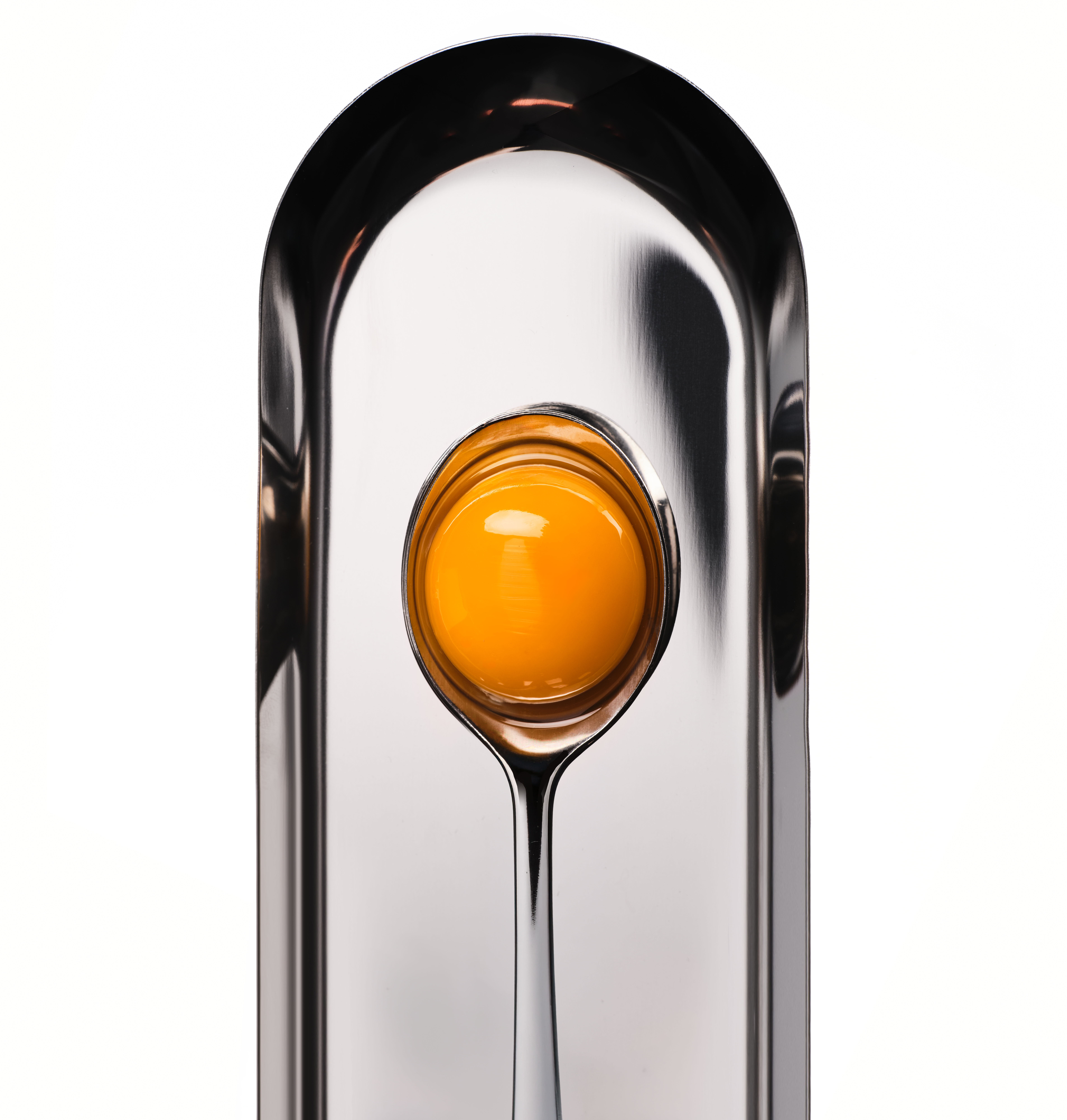 An egg yolk sits in a silver spoon, which lies on a reflective, arched silver tray against a white background.