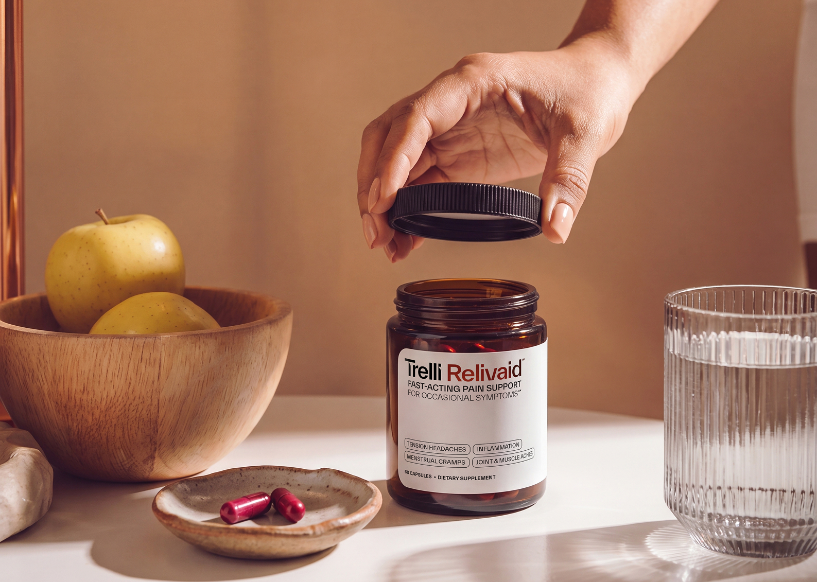 A hand opens a bottle of Trelli Relivaid, with two capsules, a glass of water, and apples nearby.