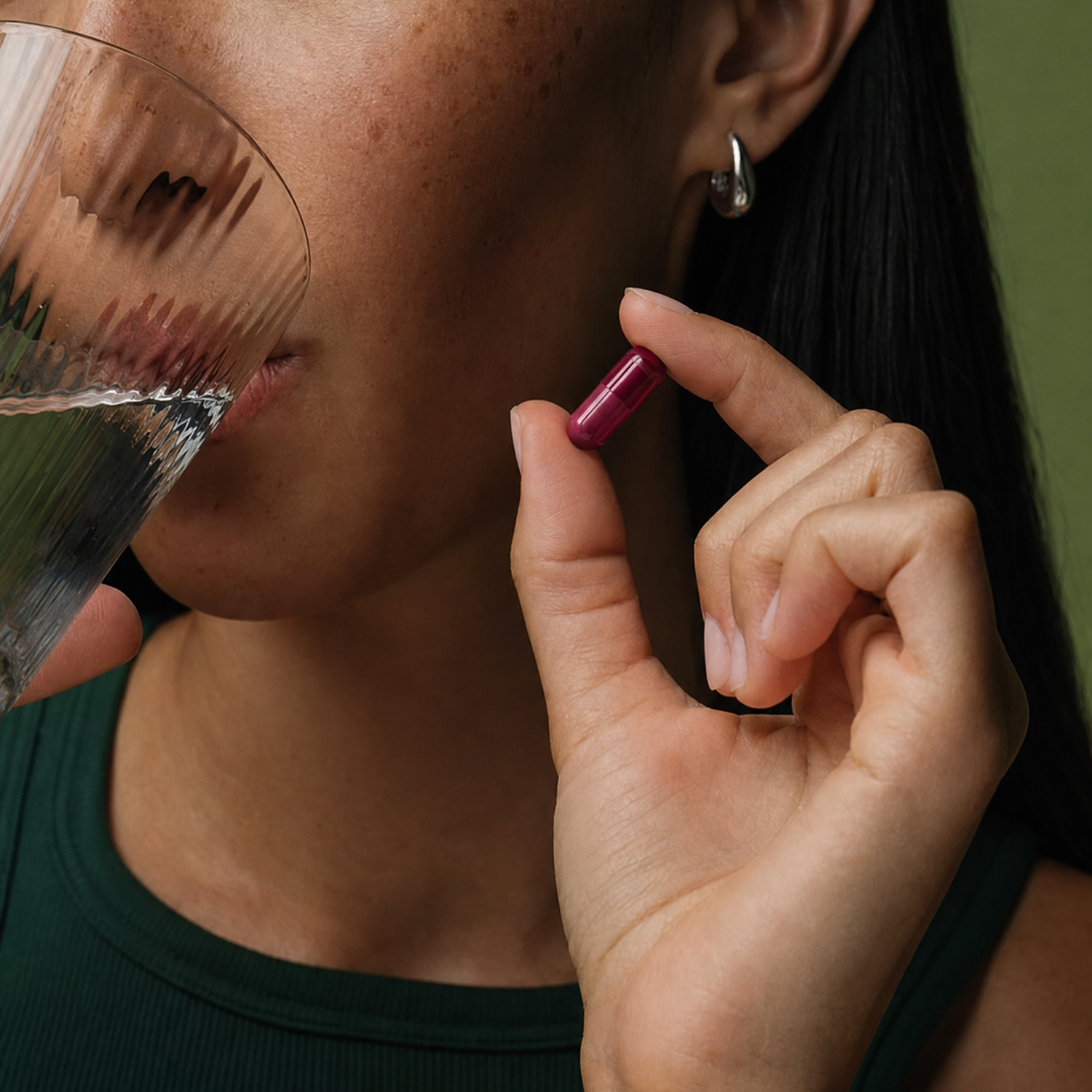 A close-up of a person holding a magenta capsule and a glass of water to their mouth.