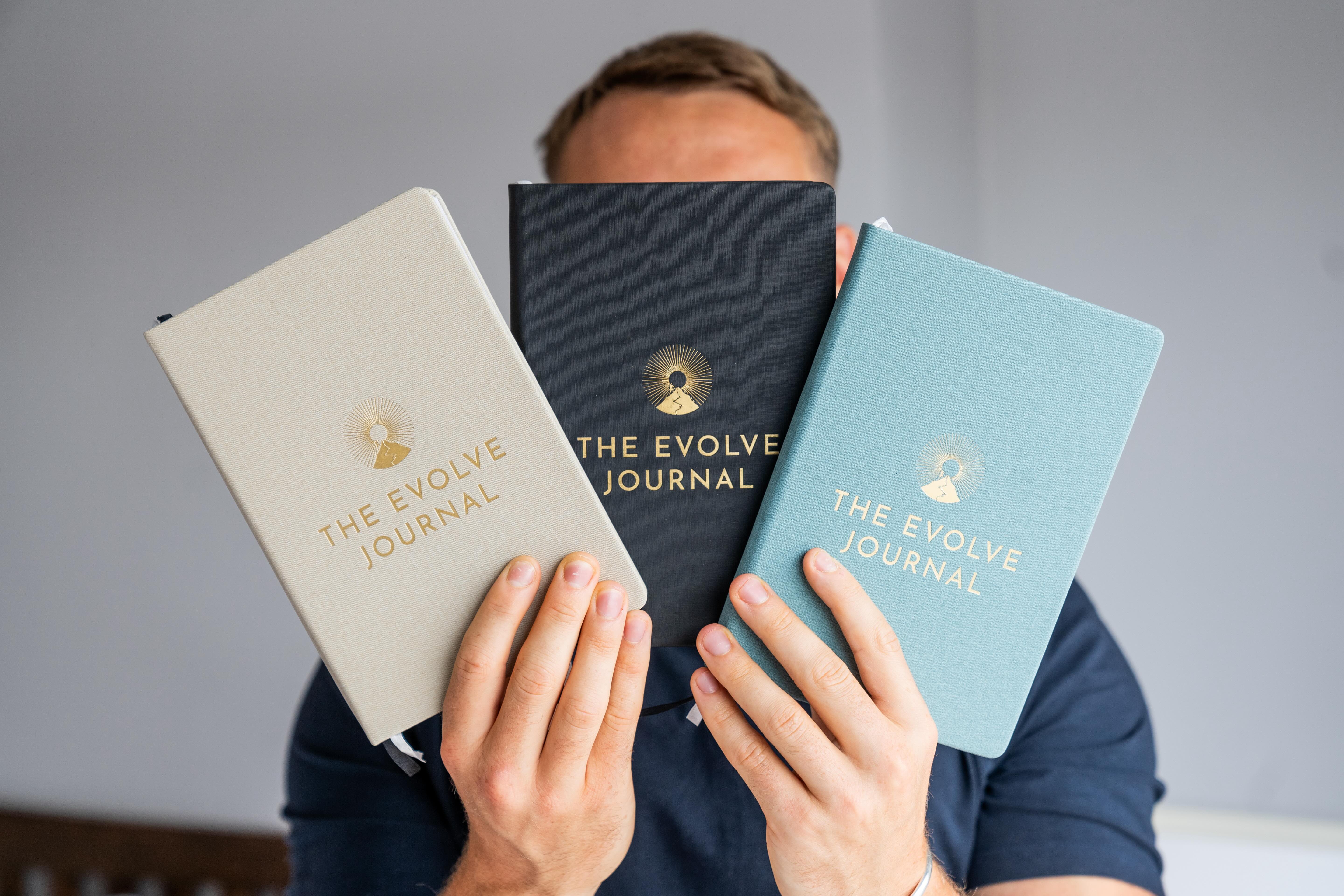 Person holding three 'The Evolve Journal' notebooks in different colors.