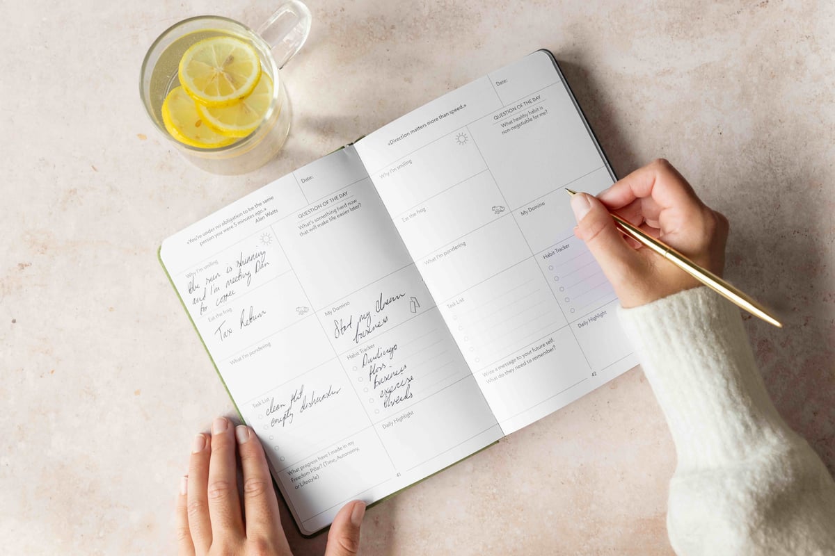 Hand writing with a gold pen in an open Evolve Journal beside a glass of lemon water
