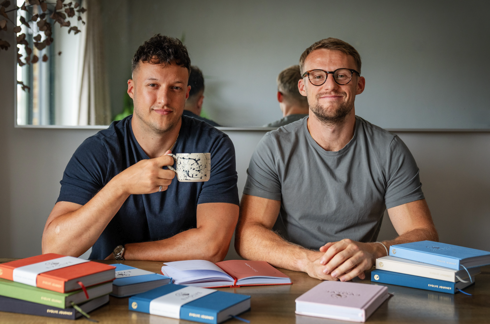 Two men sitting at a table with notebooks and a mug.