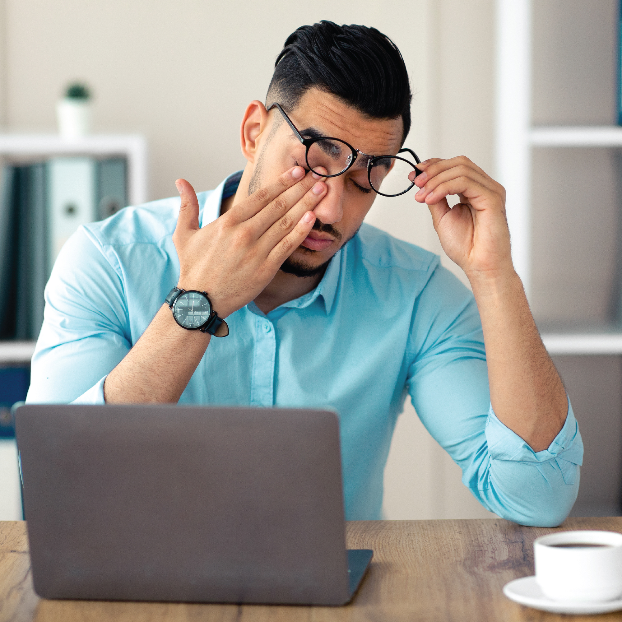 Man in blue shirt rubbing his eye at a desk with a laptop.